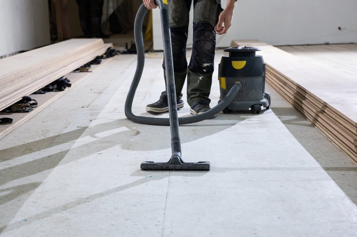A worker uses a vacuum cleaner to clean a floor before installing wooden floorboards.