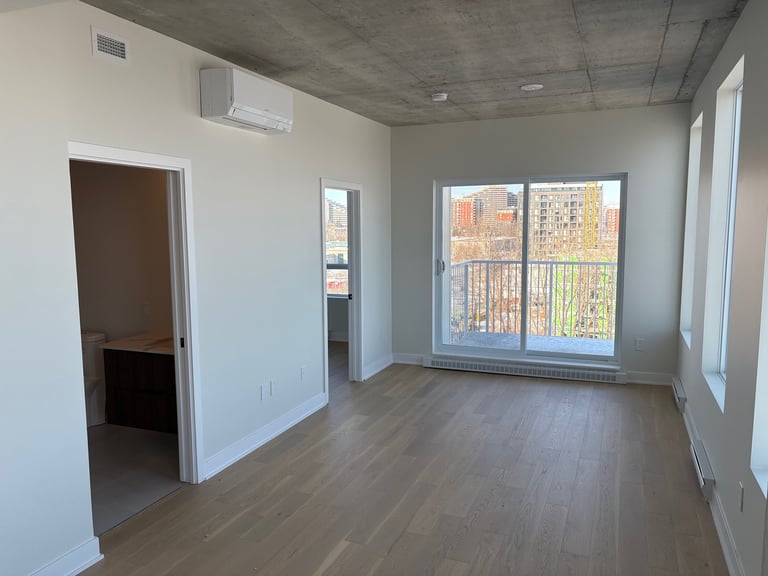 Empty apartment interior with wooden flooring, white walls, large windows, and doorway to another room