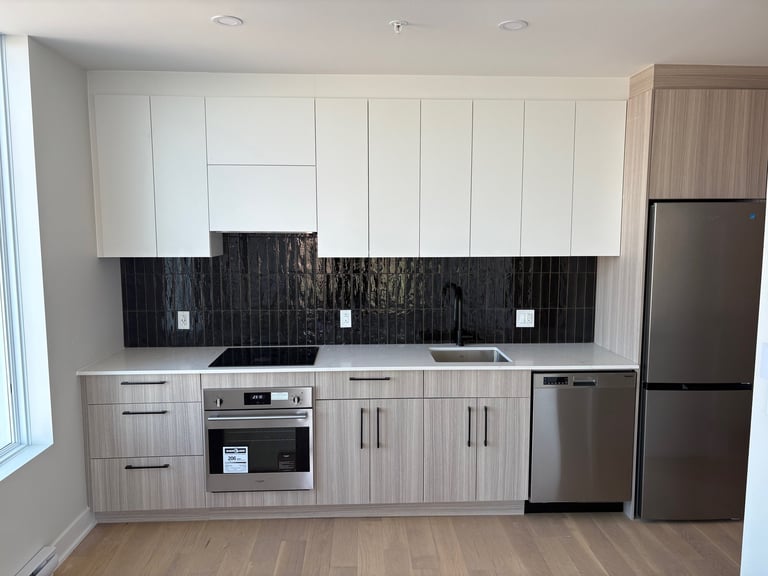 Modern kitchen with white upper cabinets, light wood lower cabinets, black tile backsplash, stainless steel appliances, and wood flooring